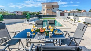 A poolside area with a table set for two, surrounded by lounge chairs, and a view of a building in the background. - Beautiful apartment in Vir (Vir)