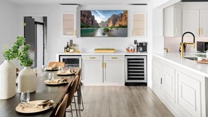 A modern kitchen with white cabinetry, a large flat-screen TV mounted above the island, and a dining area with a wooden table set for four. - Backyard Arcade | Mini Golf, Pool & Hot Tub (Scottsdale)