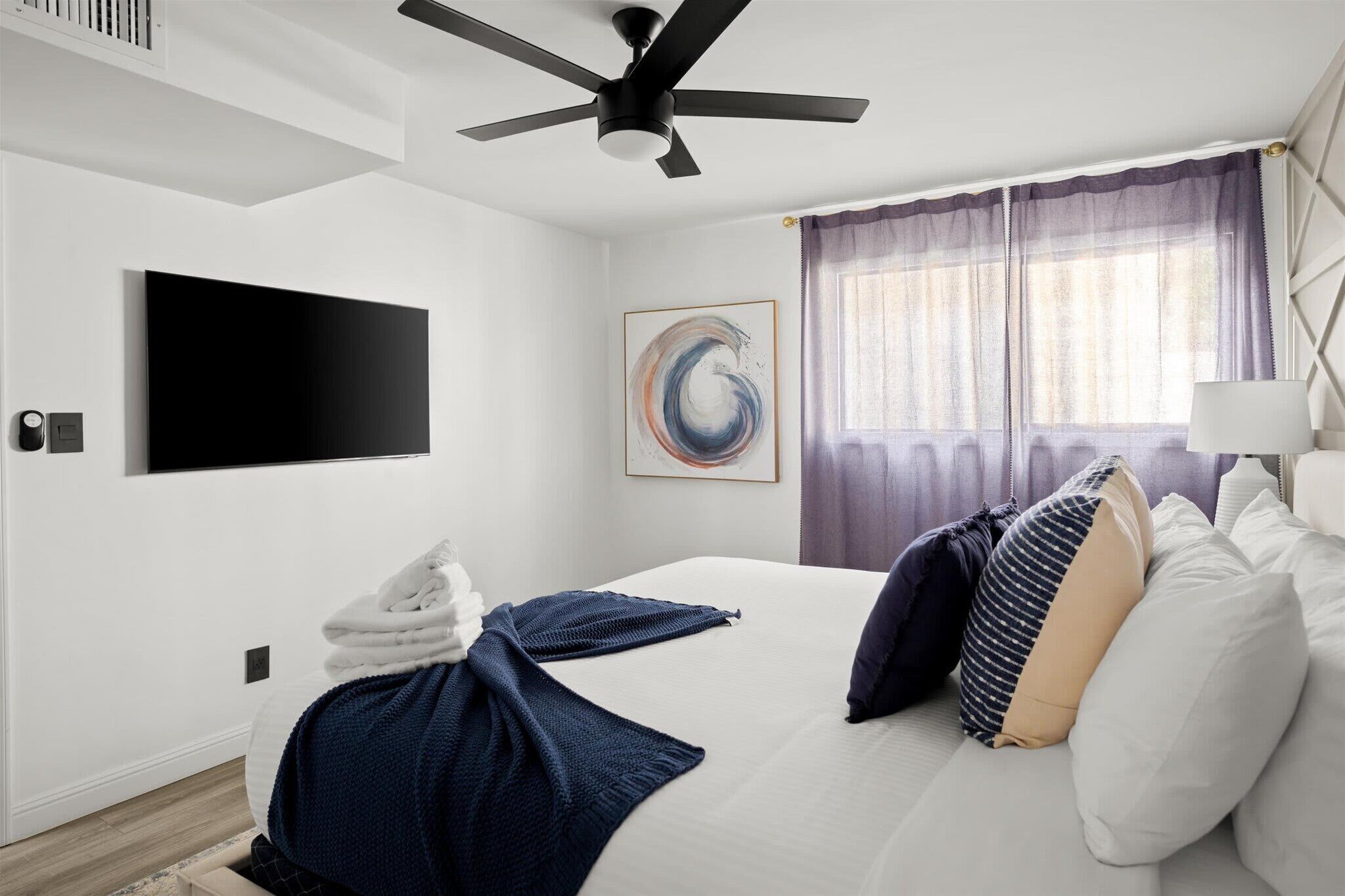 A hotel room with a bed, a ceiling fan, a flat-screen TV, a painting, and a window with curtains.