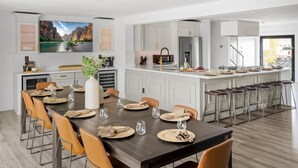 A modern kitchen with white cabinetry, a large island with bar stools, and a dining area with a long table set for six. - Backyard Arcade | Mini Golf, Pool & Hot Tub (Scottsdale)