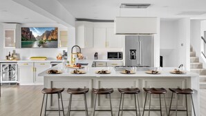 A modern kitchen with white cabinetry, a large island with bar stools, a built-in oven, and a flat-screen TV mounted above the island. - Backyard Arcade | Mini Golf, Pool & Hot Tub (Scottsdale)