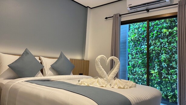 A modern hotel room with a large bed, grey pillows, a heart-shaped towel, and a view of lush greenery through a sliding glass door.