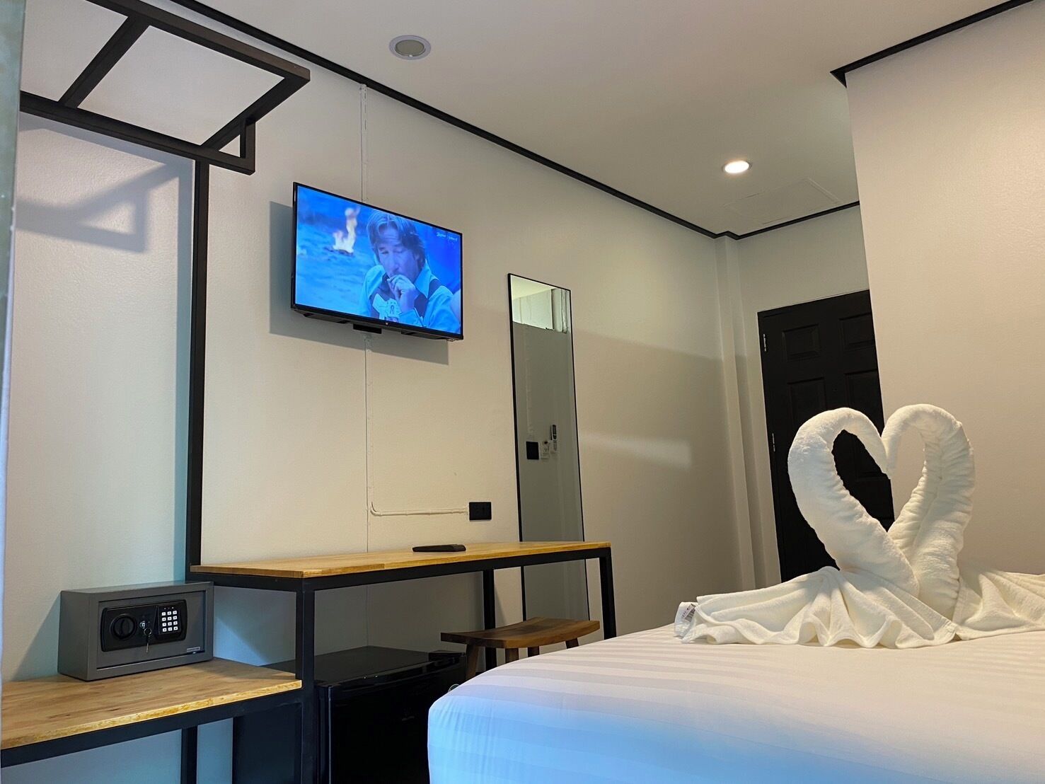 A hotel room with a bed, a desk, a TV, a safe, and a heart-shaped towel on the bed.