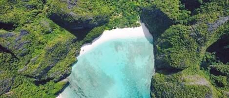 Vista aérea de una playa de arena blanca rodeada de abundante follaje verde y escarpados acantilados.