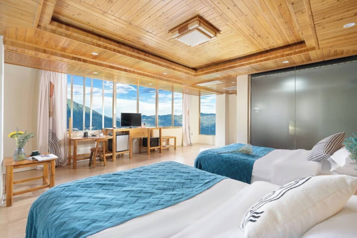 A modern hotel room with wooden ceilings, large windows offering mountain views, two beds, a desk, and a television.
