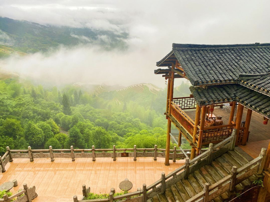 A traditional wooden pavilion with a tiled roof overlooking a lush green valley and misty mountains.