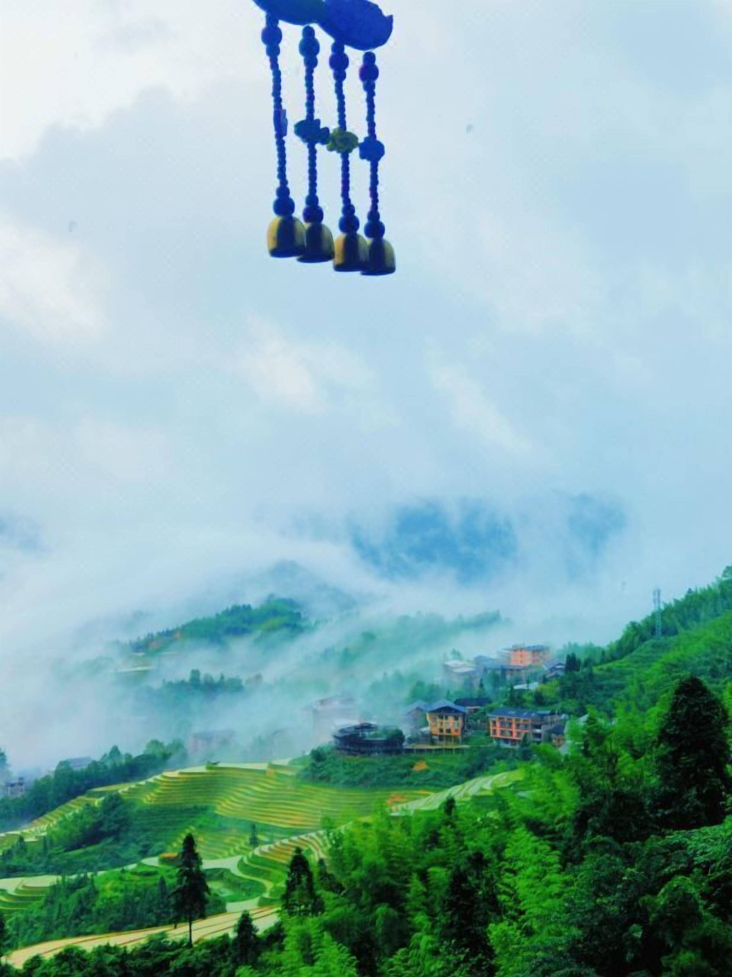 A rural landscape with terraced fields, a few buildings, and a hanging wind chime.