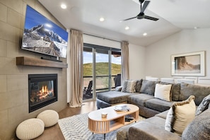 A modern living room with a fireplace, a large flat-screen TV, a sofa set, and a balcony with a view.