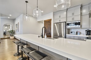 A modern kitchen with a long island, stainless steel appliances, pendant lighting, and a wooden door.