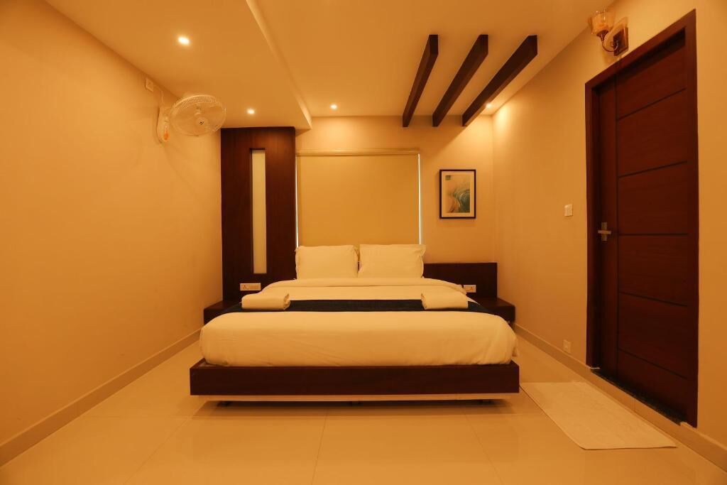 Un dormitorio con cama, mesita de noche, ventilador de pared, puerta y un cuadro enmarcado en la pared.