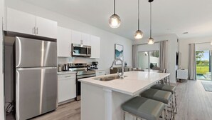 A modern kitchen with stainless steel appliances, a central island with bar stools, pendant lighting, and a view to the outdoors. - Boutique Retreat, Azzure Resort, Near Orlando and Disney by Shine Villas (Davenport)