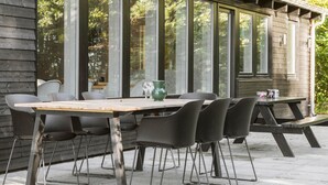 An outdoor dining area with a wooden table and metal chairs, large windows, and a view of trees.