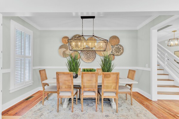 A dining area with a white table, four wicker chairs, a hanging light fixture, and decorative wall art.