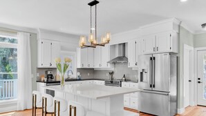 A modern kitchen with white cabinetry, a large island with bar stools, stainless steel appliances, and a hanging light fixture. - Private Pool + Fire Pit + Foosball | Short Walk to Beach | Southern Oasis (Isle Of Palms)