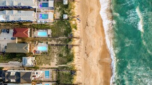 An aerial view of a coastal property with multiple buildings, including a large house with a swimming pool, smaller houses with pools, and a beach area with umbrellas. - Beachfront w/ Pool, Hot Tub & Game Room, Sleeps 28 | Atlantic Watch by AvantStay (Kill Devil Hills)