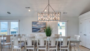 A dining area with a large table and chairs, a hanging light fixture, and a painting of a beach scene. - Beachfront w/ Pool, Hot Tub & Game Room, Sleeps 28 | Atlantic Watch by AvantStay (Kill Devil Hills)