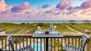 A wooden deck with a table set for two, overlooking a beach with a clear view of the ocean and dunes. - Beachfront w/ Pool, Hot Tub & Game Room, Sleeps 28 | Atlantic Watch by AvantStay (Kill Devil Hills)