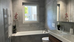 A modern bathroom with a bathtub, toilet, and sink. There is a window providing natural light and a view of the outside.