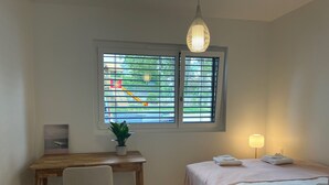 A bedroom with a bed, bedside table, lamp, desk, chair, window with blinds, and a potted plant.