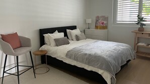 A bedroom with a bed, a chair, a nightstand, a lamp, a window with blinds, and a hanging light fixture.
