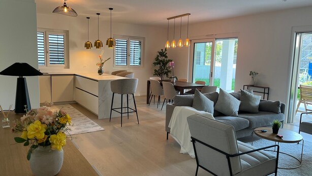 A modern kitchen with a central island, bar stools, and pendant lighting. There is a dining area with a table and chairs, and a living room with a sofa and a coffee table.