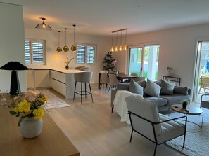 Une cuisine moderne dotée d’un îlot central, de tabourets de bar et de luminaires suspendus. On y trouve également un coin repas avec une table et des chaises, ainsi qu’un salon avec un canapé et une table basse.