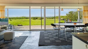 Una cocina moderna con una isla grande, un comedor con mesa y sillas, y vistas a un paisaje verde a través de puertas de cristal correderas.