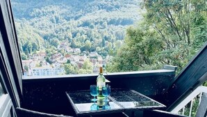 Un balcón con sillas de mimbre y una mesa, con vistas a un paisaje boscoso y casas a lo lejos.