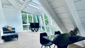 Una habitación amplia con techo alto, ventanas grandes, zona de comedor con mesa y sillas, y un sofá.