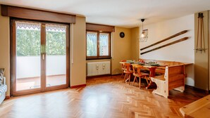 A spacious room with a wooden herringbone floor, a dining area with wooden chairs and a table, a sliding glass door leading to an outdoor area, and a ski rack on the wall. - Enrosadira by Interhome (Canazei)