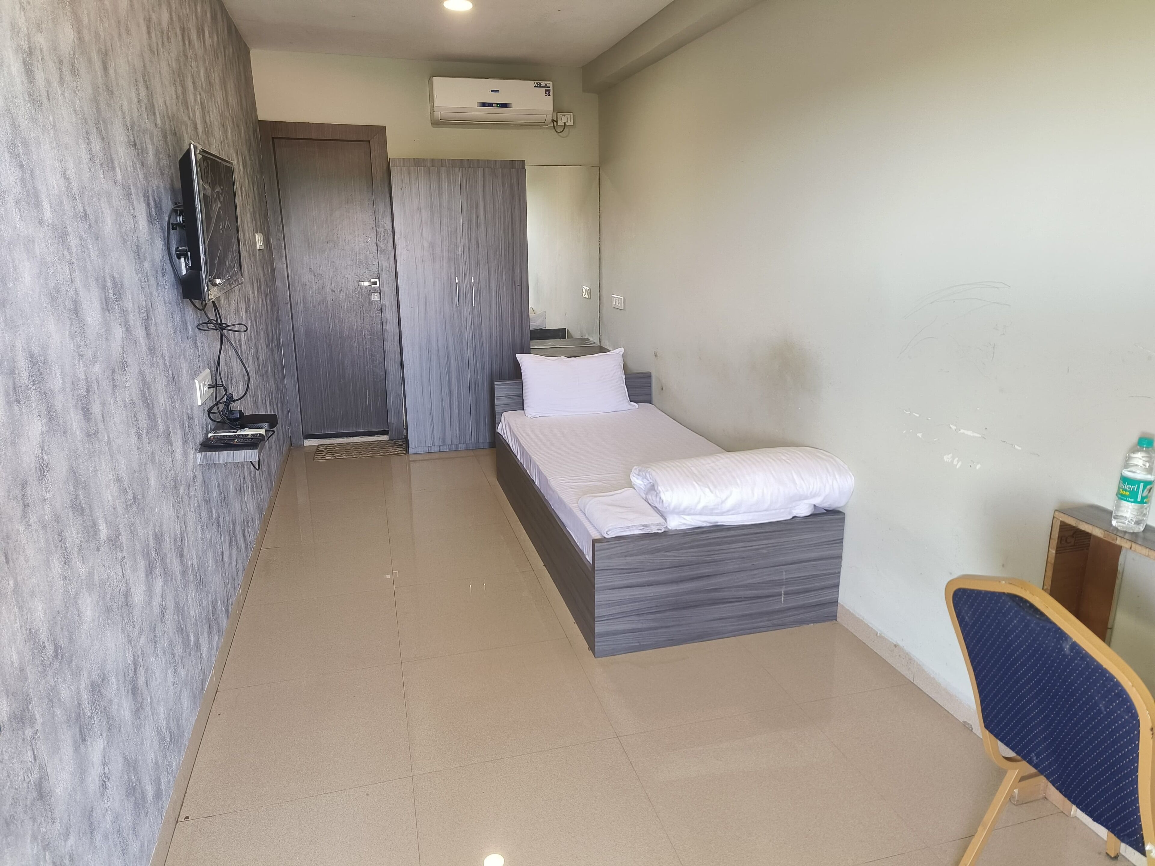 A hotel room with a bed, a chair, a TV mounted on the wall, and an air conditioning unit.