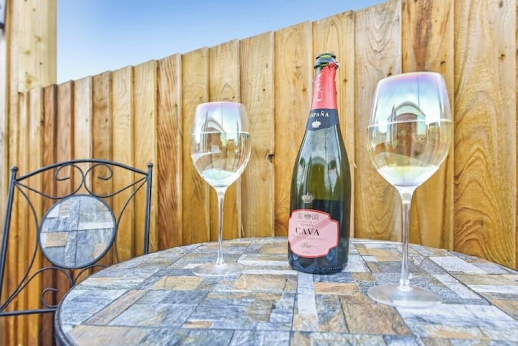 Een tafel met een fles cava wijn en twee glaasjes, voor een houten omheining.