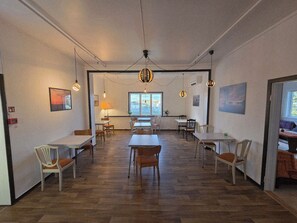 A spacious dining area with wooden flooring, multiple tables and chairs, pendant lighting, and a large window providing natural light. - Våg Helgeland (Alstahaug)