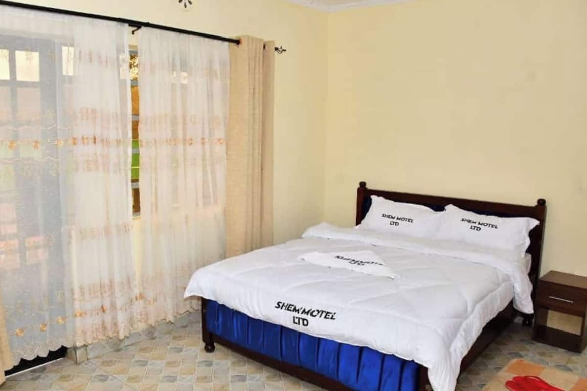 A single bed with a white bedspread and blue skirted bed base, a wooden headboard, and a nightstand with a lamp.