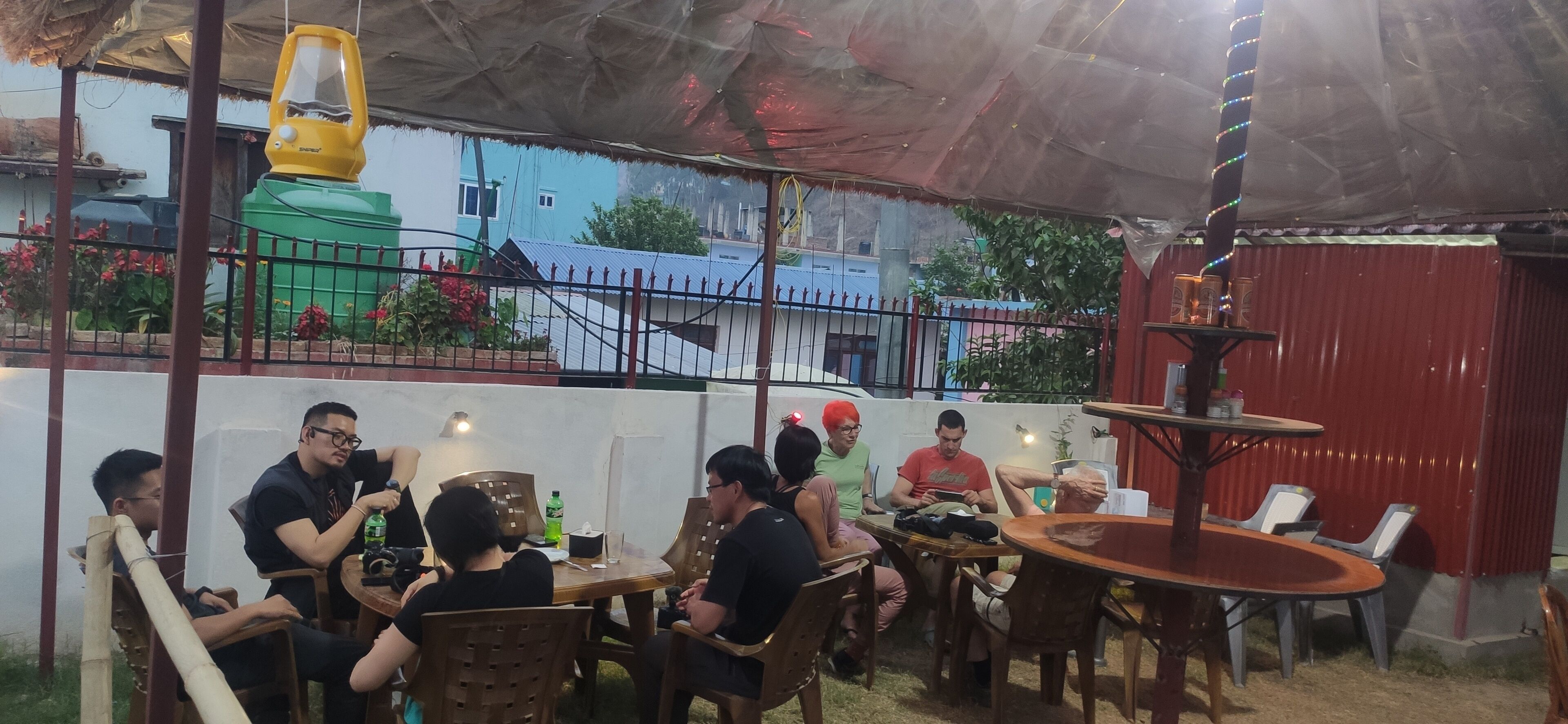 A group of people seated at tables under a covered patio area with a view of buildings and a fence.