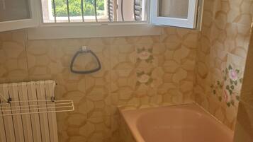 A bathroom with a bathtub, a toilet, a window with a view of greenery, and patterned walls.