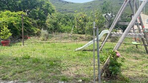 Un mur de pierres, une balançoire en bois et un toboggan en métal, le tout installé dans un espace verdoyant, avec des arbres et des collines en arrière-plan.