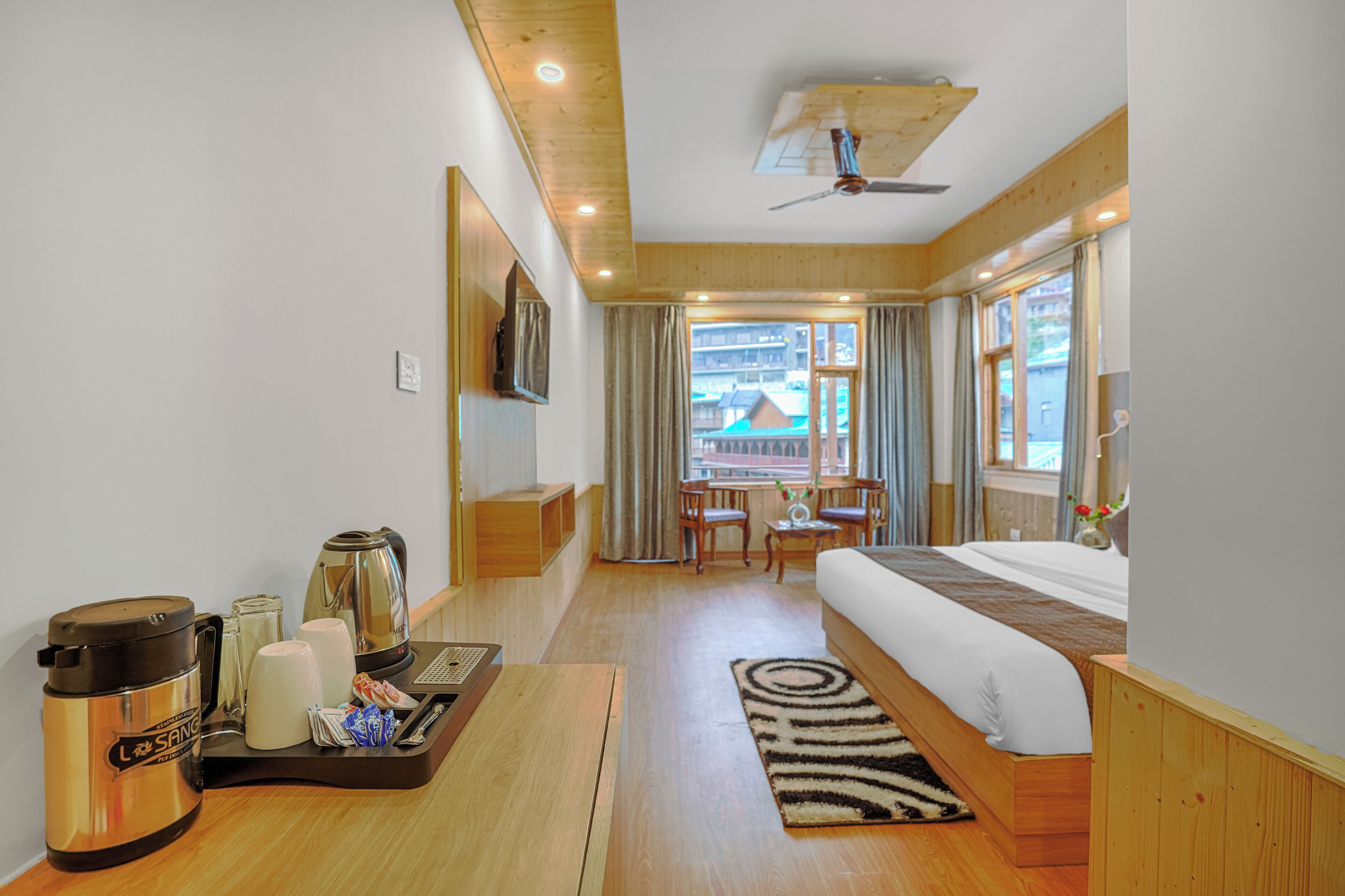 Habitación de hotel con cama, escritorio con tetera, cafetera y bandeja con tazas y tetera. Se ve una ventana con cortinas y vistas a edificios.