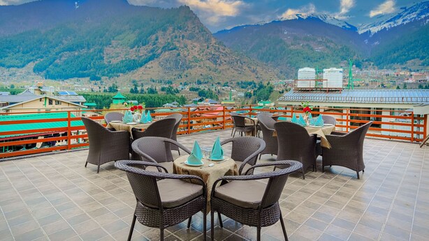 A terrace with wicker furniture and tables, overlooking a mountainous landscape with a town in the background. - Hotel Divine Paradise Manali (Manali)