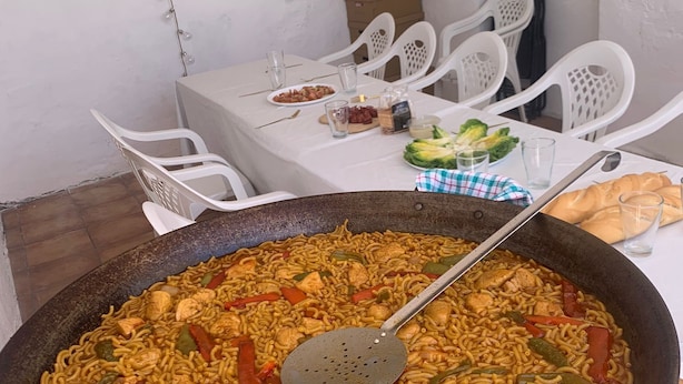 Ein Esstisch mit einer großen Pfanne gebratener Nudeln und Gemüse, weißen Plastikstühlen und einer weißen Tischdecke.
