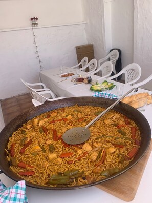 A dining table with a large pan of stir-fried noodles and vegetables, white plastic chairs, and a white tablecloth. - Peace and Quiet (Lillo)