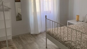 Ein Schlafzimmer mit Bett, Nachttisch, Lampe, Kleiderschrank und Fenster mit leichtem Vorhang.