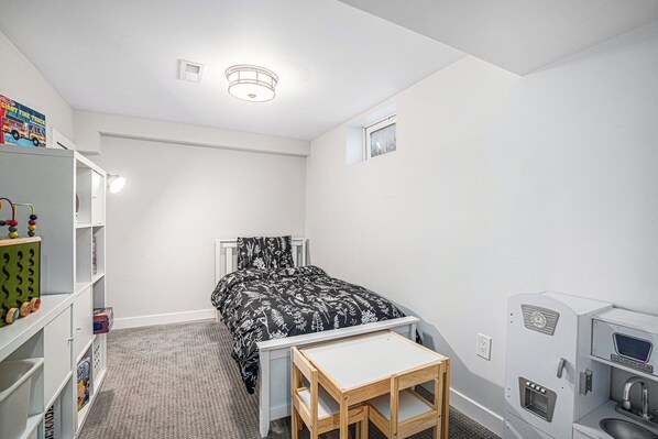 A child's bedroom with a bed, a small table, a toy shelf, and a kitchenette.