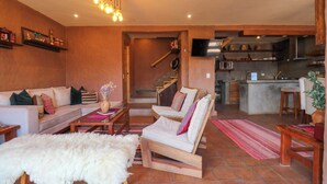 A cozy living area with a sofa, a wooden chair, and a fur rug. A staircase leads to a kitchen area with a TV and a sink. - w* | Splendid 3BR House in Valle Sagrado (Urubamba)