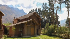 Une maison rustique en bois, avec un toit de chaume, entourée d’une pelouse bien entretenue et de grands arbres.