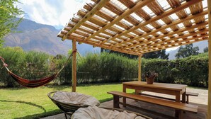 En træpergola med et picnicbord og to polstrede stole i en have med bjergudsigt.