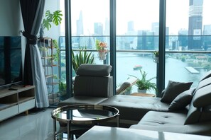 A modern living room with a sofa, a coffee table, and a TV. There are plants on a balcony railing and a city view through large windows. - Metropole - Christine Hospitality (Ho Chi Minh City)