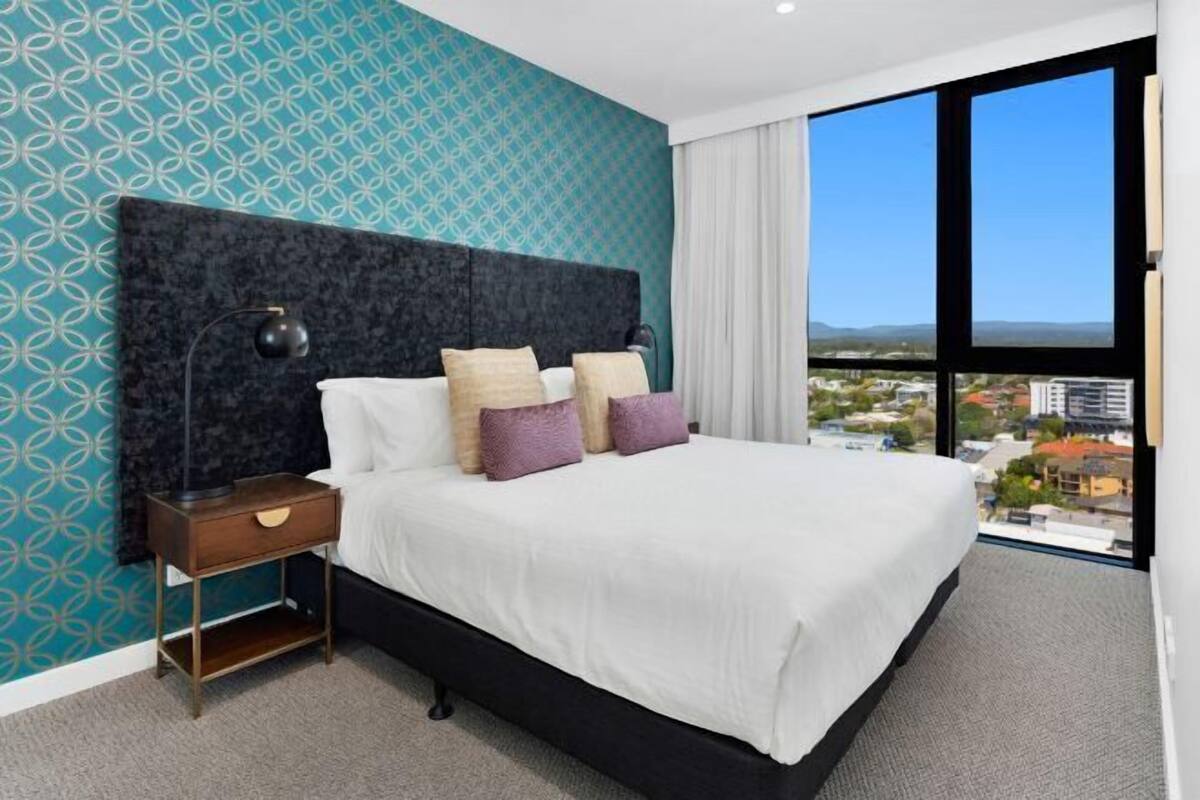 A hotel room with a large bed, a nightstand, a window with a city view, and patterned wallpaper.