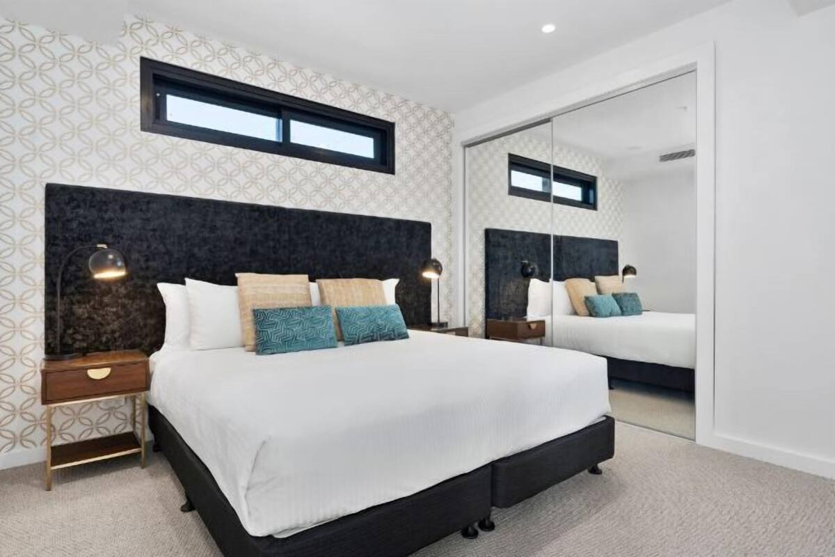 A modern bedroom with a large bed, two bedside tables, a nightstand, a large mirror, and a window.