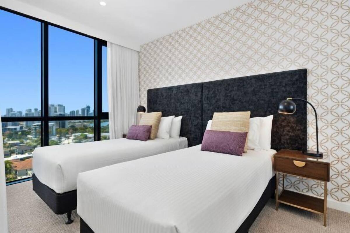 A hotel room with two beds, a large window with a city view, a patterned wall, and a nightstand with a lamp.